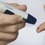Person holding a lancing device next to a fingertip with a small drop of blood for a home blood sugar check
