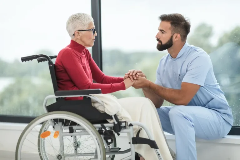 Nurse kneeling beside an older patient and holding her hands during a care conversation