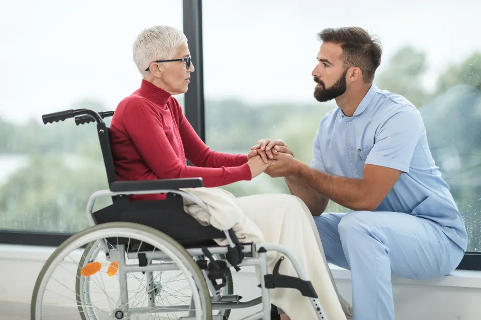 Nurse kneeling beside an older patient and holding her hands during a care conversation